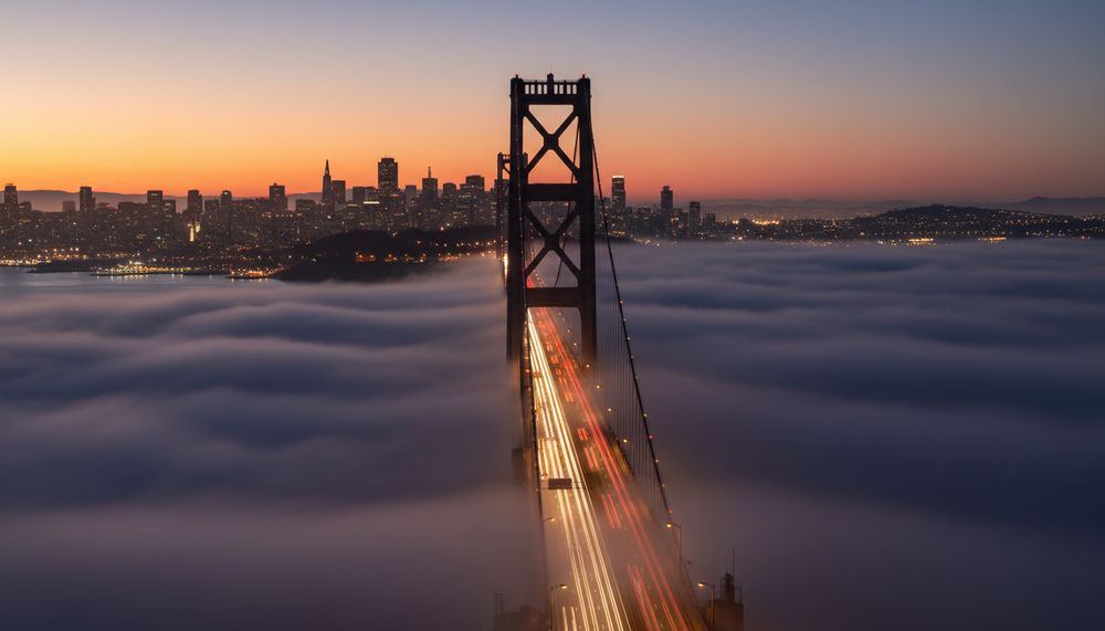 The Golden Gate Bridge rising out of a low fog bank at dusk with the lit San Francisco skyline behind it